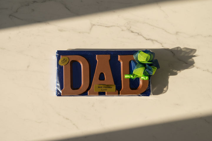 A person holding a large chocolate gift box with the word 'DAD' in brown letters, wrapped in shiny blue foil with a green bow, in a store setting.